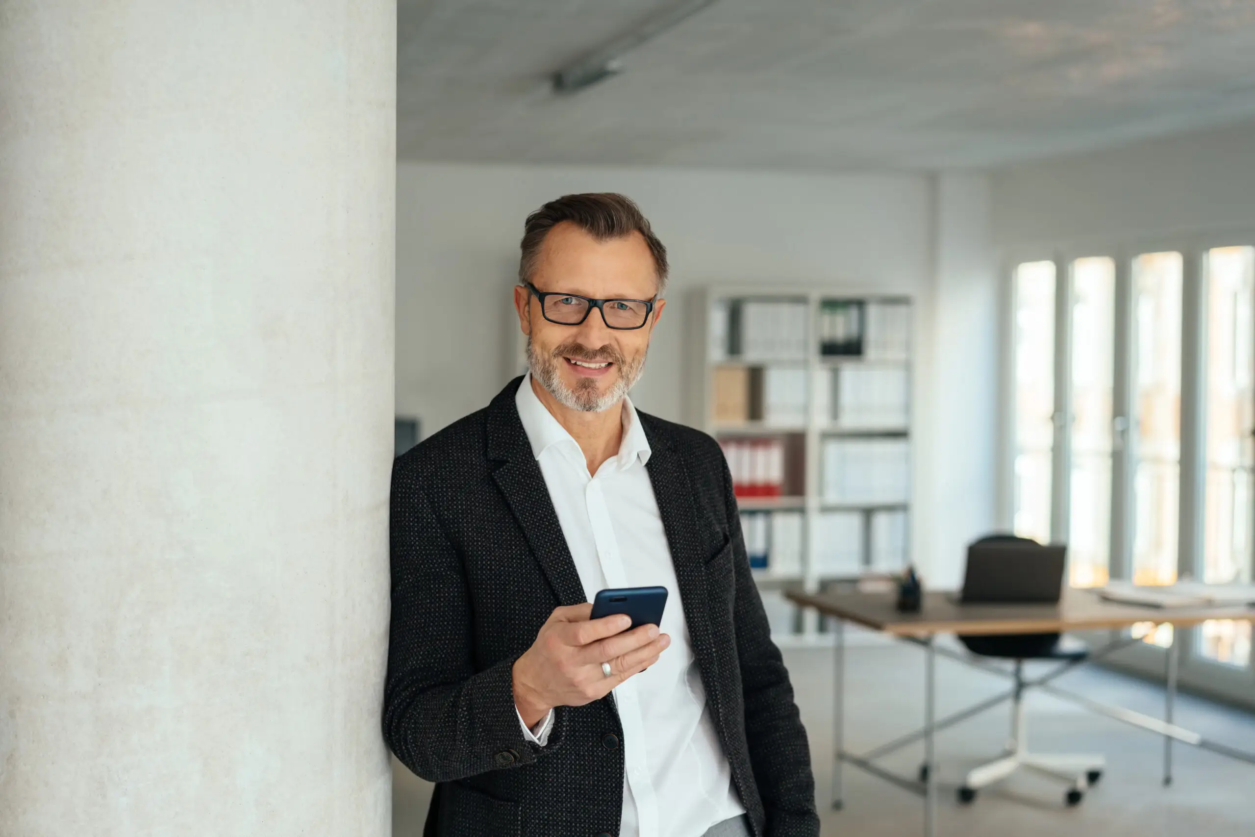 Stylish businessman holding his mobile phone as he relaxes leaning against an interior office wall smiling at the camera