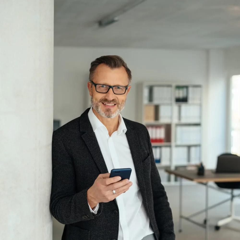 Stylish businessman holding his mobile phone as he relaxes leaning against an interior office wall smiling at the camera